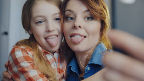 Portrait-shot-of-the-beautiful-woman-with-young-daughter-making-selfies-on-the-smart-phone,-showing-their-tongues,-hugging-and-laughing.-Close-up.-Inside